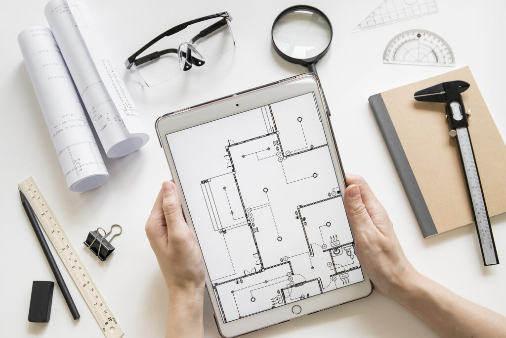 Pessoa segurando um tablet que exibe uma planta baixa digital, com instrumentos de desenho ao redor.