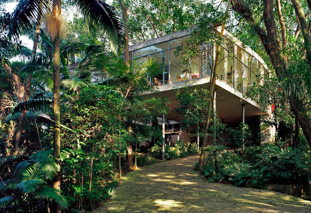 View of the iconic Glass House in São Paulo, Brazil, surrounded by lush tropical vegetation. The modernist residence, designed by architect Lina Bo Bardi, features floor-to-ceiling glass walls elevated on slender stilts above the forested landscape.