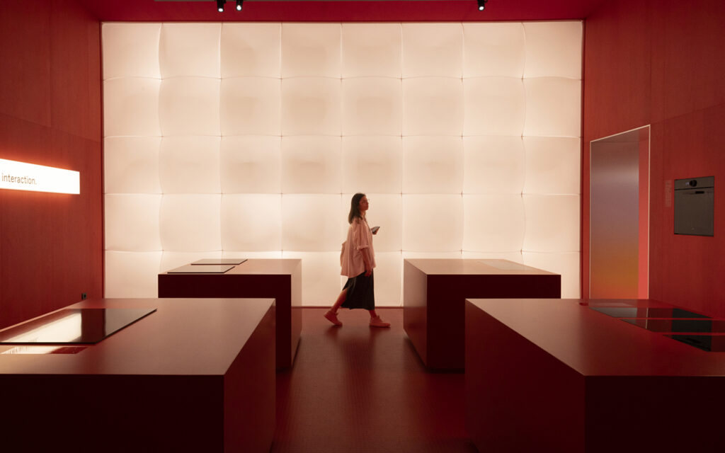 Mulher em estande moderno com balcões minimalistas vermelhos e parede iluminada, representando tendências de design de interiores e inovação tecnológica esperadas no Salão de Milão 2026.